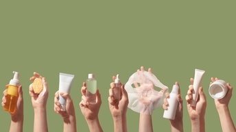 Vor grünem Hintergrund sind dreizehn Hände in die Luft gestreckt. Jede Hand hält ein anderes Pflegeprodukt, zum Beispiel eine Tube, einen Schwamm, eine Gesichtsmaske, eine Dose, eine Pumpflasche etc