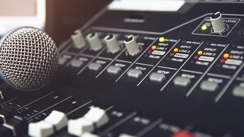 Nahaufnahme eines Mischpults, auf dem ein paar Leuchtkontrollen orange oder gelb aufleuchten. Auf dem Mischpult liegt ein Mikrofon