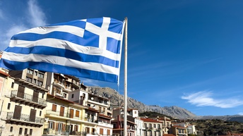 Die griechische Flagge weht im Wind, im Hintergrund eine Stadtansicht.
