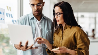 Zwei Personen stehen in einem Raum dicht nebeneinander. Die linke Person hält mit der rechten Hand einen aufgeklappten Laptop. Auf den Bildschirm des Laptops deutet die rechte Person mit einem länglichen Gegenstand