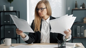 Person mit Brillen sitzt an Tisch und sichtet Dokumente ,weitere Unterlagen und Tasse auf Tisch platziert, im Hintergrund verschwommen Regale mit Ordnern