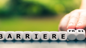 Weiße Holzwürfel nebeneinander stehend mit schwarzer Aufschrift barrierefrei, die letzten beiden Würfel werden von einer Hand nach hinten gekippt mit dem Wort frei