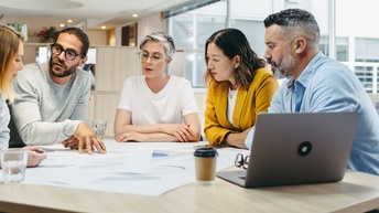 Fünf Personen sitzen um einen Tisch. In der Mitte auf dem Tisch liegt ein großer Zettel, auf den die Personen teilweise blicken. Eine Person deutet darauf. Neben dem Zettel steht ein aufgeklappter Laptop