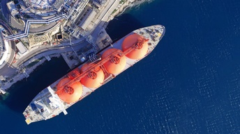 Top-Shot eines großen, langen Schiffes in einem blauen Gewässer. Auf dem Schiff sind vier große, orange Silos. Das Schiff ist direkt neben einer asphaltierten Oberfläche, auf der ein große Anlage steht.