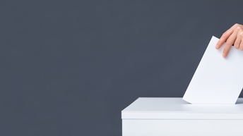 Seitliche Aufnahme der Hand einer Person, die ein Briefkuvert in einen weiße Box wirft. Der Hintergrund ist grau
