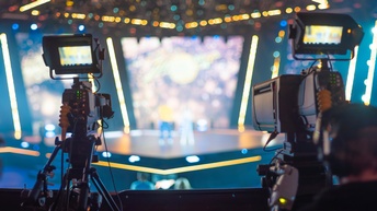 Zwei verschwommene Köpfe von Personen, die Hinter Filmkameras stehen, die Kameras auf große Showbühne gerichtet