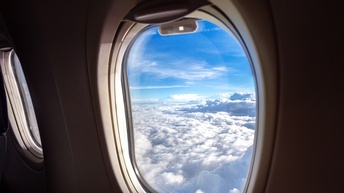Ausblick aus einem Flugzeugfenster auf einen hellblauen Wolkenhimmel.