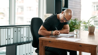 Eine schwarz gekleidete Person mit Brille sitzt in einem Büro und schreibt fokussiert.
