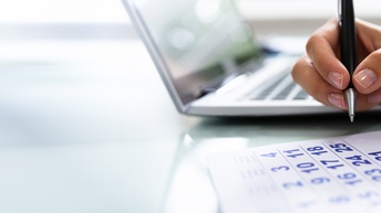 Nahaufnahme der Hand einer Person, die einen Kugelschreiber hält. Unter der Hand ist ein Kalenderblatt. Daneben steht ein aufgeklappter Laptop