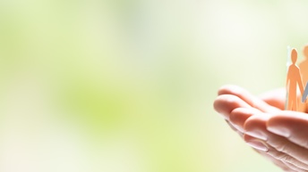 In der rechten Bildhälfte sind zwei nach oben gewandte Hände. Auf den Händen sind kleine Papierfiguren von Personen, die sich an den Händen reichen und einen Kreis bilden