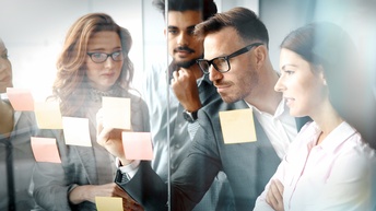 Gruppe von unterschiedlichen Personen steht vor einer Glaswand mit Post-Its und betrachtet diese