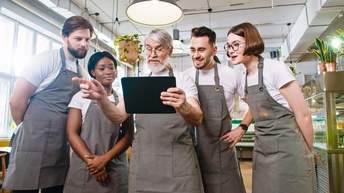 Frontale Aufnahme von fünf Personen mit Schürzen, die nebeneinander aufgereiht in einem Lokal stehen. Die mittlere Person hält ein Tablet, auf das alle blicken.