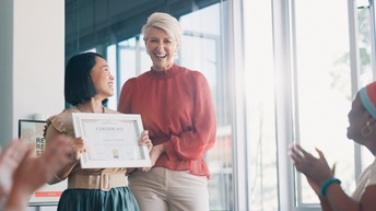 Zwei Personen stehen nebeneinander. Die linke Person hält einen Bilderrahmen, in dem ein Zettel ist, auf dem Zertifikat steht. Den beiden Personen gegenüber sind zwei Personen, die klatschen
