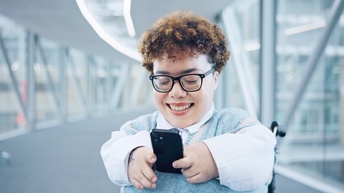 Lächelnde Person mit Brillen befindet sich in gläsernem Gang und blickt auf Smartphone