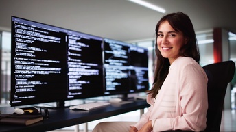 Porträt einer Person mit offenen, braunen Haaren, die an einem Schreibtisch sitzt und lächelt. Vor ihr auf dem Schreibtisch sind vier Bildschirme. Auf jedem Bildschirm ist ein HTMl-Code