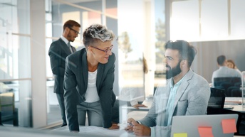 Zwei Personen mit kurzen Haaren und Businesskleidung unterhalten sich bei einem Büroarbeitsplatz, im Hintergrund zeigen sich weitere Arbeitsplätze in einem hellen Großraumbüro mit weiteren Personen