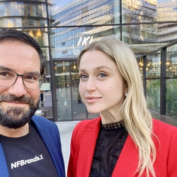 Porträt von zwei nebeneinander stehenden Personen vor einem Gebäude auf einem asphaltierten Platz. Die linke Person hat kurze, dunkle Haare und trägt Brille. Die rechte Person hat offene, helle Haare und trägt ein dunkles Oberteil