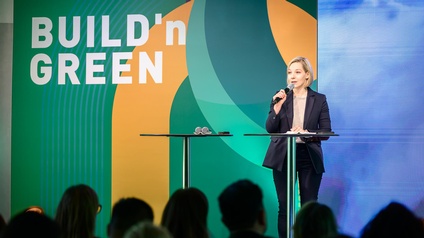 Eine Person steht auf einer Bühne hinter einem Stehtisch und spricht in ein Mikrofon. Vor der Bühne ist ein sitzendes Publikum. Neben dem Stehtisch befindet sich ein weiterer Tisch mit Mikrofonen. Im Hintergrund ist eine großflächige, farbige Bühnenwand mit den Worten „BUILD’n GREEN“