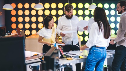 Fünf Personen stehen in einem Büro um einen Tisch. Zwei der Personen halten jeweils einen Zettel. Auf dem Tisch liegen ein Tablet, Post-its, ein Laptop sowie andere Büroutensilien. Die Personen reden miteinander