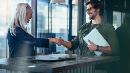 Zwei lächelnde Personen reichen sich über Stehtisch die Hand