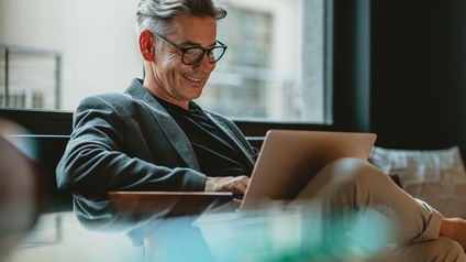 Person mit kurzen grauen Haaren und Brille sitzt mit einem Laptop auf dem Schoß und blickt freudig darauf
