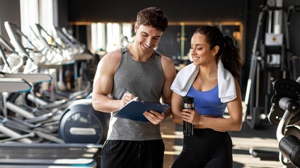 Zwei Personen in Sportkleidung stehen nebeneinander. Die linke Person notiert etwas auf einem Klemmbrett, auf das beide blicken. Die rechte Person hat ein Handtuch um den Hals. Im Hintergrund stehen verschiedene Sportgeräte