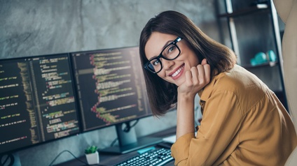Lächelnde Person mit Brillen stützt Kopf in die Hand an Schreibtisch mit Monitoren, auf denen Quellcodes zu sehen sind, sitzend