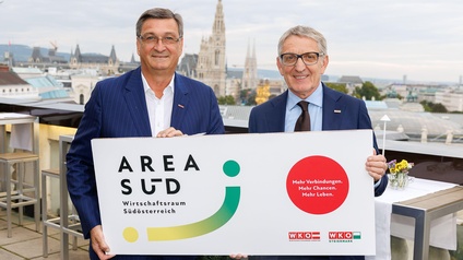 Mandl und Herk halten eine Tafel auf einer Dachterasse - im Hintergrund das Rathaus Wien