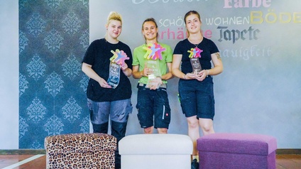 Die drei Siegerinnen des Landeslehrlingswettbwerbs mit Trophäe in der Hand. Im Vordergrund befinden sich drei frisch tapezierte Hocker.