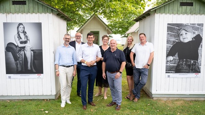 Gruppenfoto zwischen zwei weißen Badehütten aus Holz im Strandbad Klagenfurt. Auf den Hütten sind zwei große Fotos der Ausstellung der Berufsfotografen zu sehen. 