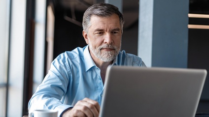 Person mit kurzen weiß-grauen Haaren und Bart mit blauem Hemd sitzt freudig vor einem Laptop in einem modernen Büroraum