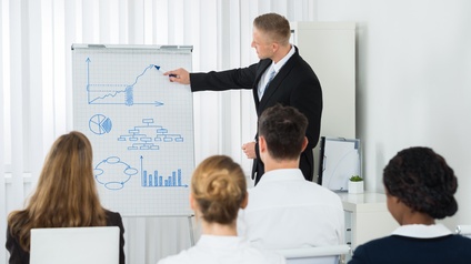 Person malt mit Stift auf Flipchart Diagramme, im Vordergrund mehrere Personen sitzend in Rückenansicht