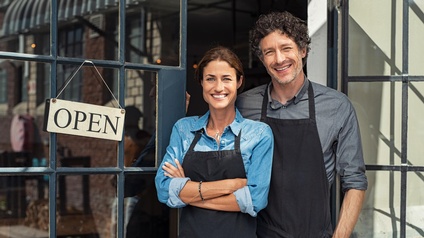 Zwei lächelnde Personen mit schwarzen schürzen stehen im Eingangsbereichs eines Geschäftes, an der Tür hängt ein Schild mit dem Schriftzug open