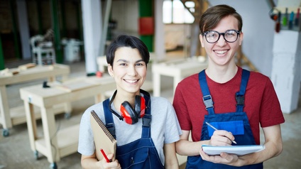 Zwei lächelnde Personen in blauen Arbeitslatzhosen mit Stiften und Notizblöcken in Tischlerwerkstatt stehend