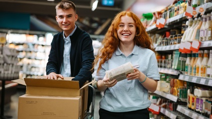 Porträt zweier nebeneinander stehender jugendlicher Personen. Eine Person greift in einen geöffneten Karton, der vor ihr erhöht auf einem Rollwagen steht. Die andere hält eine rechteckige Packung. Rechts daneben ist ein mit Lebensmitteln befülltes Regal.