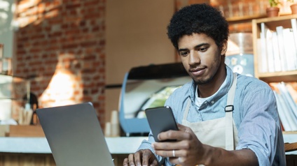 Eine Person mit Schürze sitzt an einem kleinen, runden Tisch und blickt auf ein Handy, das sie hält. Auf dem Tisch steht ein aufgeklappter Laptop. Im Hintergrund ist ein Tresen