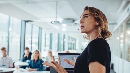 Eine Person mit einem Mikrofon vor den Lippen steht hinter einem Pult und gestikuliert mit den Händen. Auf dem Pult steht ein Laptop. Ihr gegenüber sitzen fünf Personen jeweils einzeln hinter einem Tisch