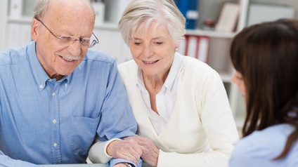 Senioren-Paar sitzt bei einem Tisch und blickt freudig auf ein Tablet, das von einer anderen Person gehalten wird, Beratungsgespräch