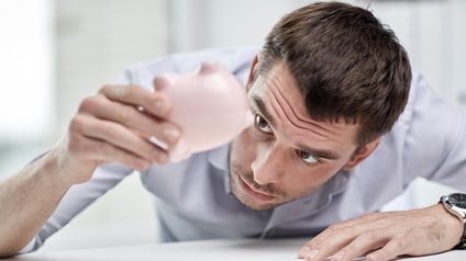 Person mit kurzen dunklen Haaren blickt erwartungsvoll auf ein ausgeleertes  Sparschwein während Münzen auf dem Tisch liegen