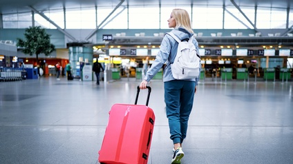 Rückenansicht einer Person, die gerade einen Flughafen betritt und ihren roten Koffer mit sich zieht. Am Rücken trägt sie einen weißen Rucksack und schaut auf die Seite.