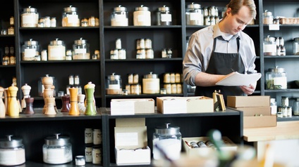 Eine Person mit Schürze steht in einem Geschäft hinter einem Tresen. In der Hand hält sie Zettel, auf die sie blickt. Auf dem Tresen stehen Pfeffermühlen. Hinter der Person sind in einem Regal verschiedene Gewürze