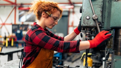 Seitenansicht einer Person, die eine industrielle Bohrmaschine bedient. Die Person trägt Arbeitskleidung und wirkt konzentriert. Im Hintergrund ist eine Industriehalle zu sehen.