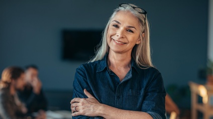 Person mittleren Alters mit langen weiß-grauen Haaren, Brille und blauem Jeanshemd steht lächelnd und mit verschränkten Armen in einem Raum während im Hintergrund andere Personen sitzen