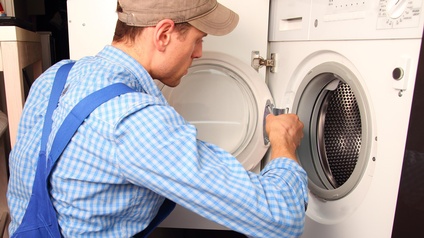 Person in blauer Arbeitskleidung schraubt am Bullauge einer Waschmaschine