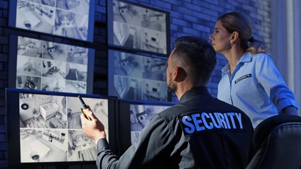 Eine sitzende Person in Rückenansicht mit dem Schriftzug Security am Hemd hält ein WalkieTalkie in Händen und blickt mit einer neben ihr stehenden Person auf sechs Bildschirme, die verschiedene Aufnahmen von Räumen zeigen.
