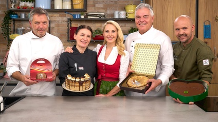 Porträt von fünf Personen, die nebeneinander an einer Küchenzeile stehen. Vier Personen tragen weiße Kochkleidung und halten eine Torte. Die fünfte Person trägt ein Dirndl. Alle lächeln