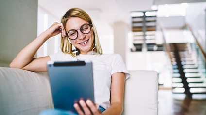Lächelnde Person mit Brillen und Kopfhörern sitzt auf einer Couch, stützt einen Arm an den Kopf, hält in anderer Hand ein Tablet, auf das sie blickt.