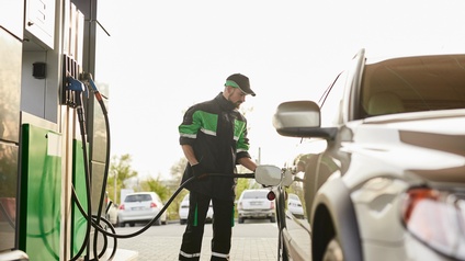Eine Person in Arbeitskleidung steht an einem Auto neben einer Zapfsäule auf eine Tankstelle und tankt das Gefährt.