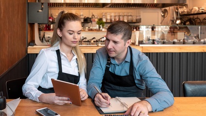Zwei Personen in Arbeitskleidung sitzen an einem Tisch in einem Restaurant und blicken auf ein Tablet während mit einem Taschenrechner gerechnet wird und mit einem Stift Notizen gemacht werden