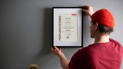 Person mit kurzen dunklen Haaren, Bart, roter Mütze und rotem Shirt steht vor einer grauen Wand und hält einen Bilderrahmen mit Passepartout und Prüfungszeugnis Meister in Händen, daneben steht eine Pflanze